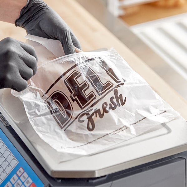 A person in gloves is putting food in a "Deli Fresh" printed plastic bag on a counter.