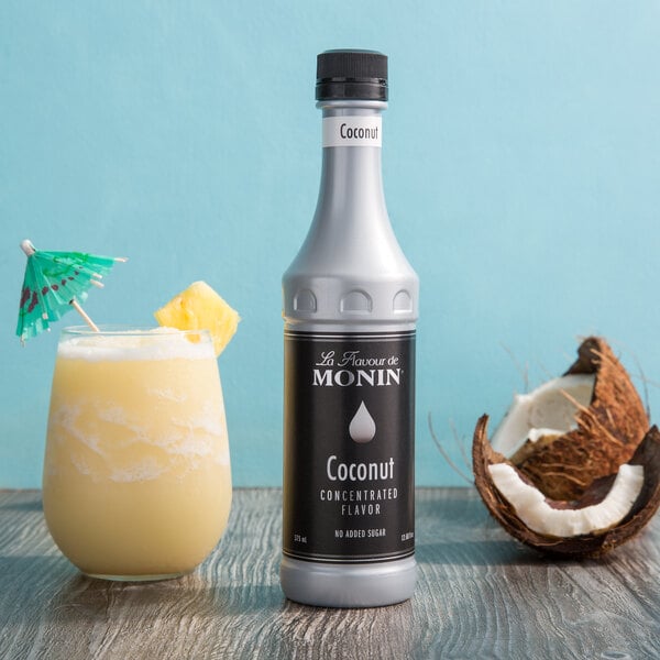 A close-up of a bottle of Monin Premium Coconut Concentrated Flavor next to a glass of yellow liquid.
