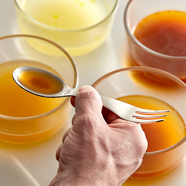 A hand holding a Mercer Taste stainless steel tasting spoon over a glass of liquid.
