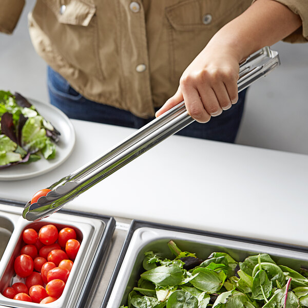 A person using Vollrath Jacob's Pride stainless steel scalloped utility tongs to serve cherry tomatoes on a salad bar tray.