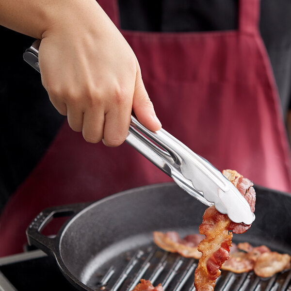 A hand holding Vollrath stainless steel utility tongs over bacon cooking in a skillet.
