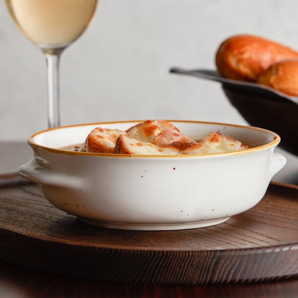 An Acopa Keystone vanilla bean stoneware onion soup crock on a wooden tray with a bowl of French onion soup and a glass of wine.