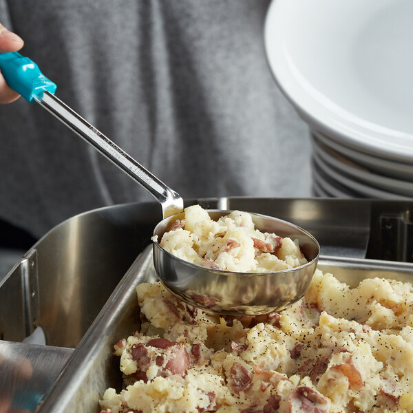 A hand using a Vollrath teal stainless steel spoodle to serve mashed potatoes from a metal pan.