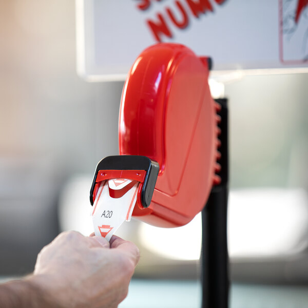 A hand holding a red 3-digit paper ticket from a Garvey Take-A-Number dispenser.