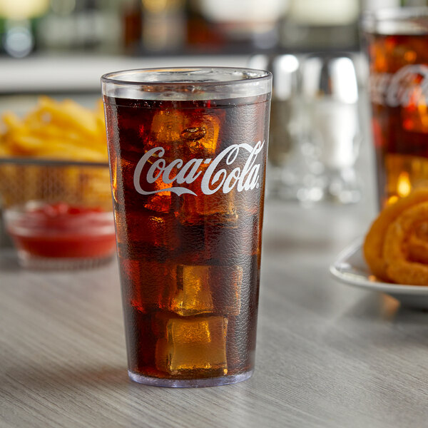 A clear plastic Coca-Cola tumbler filled with soda and ice on a table with a plate of onion rings.