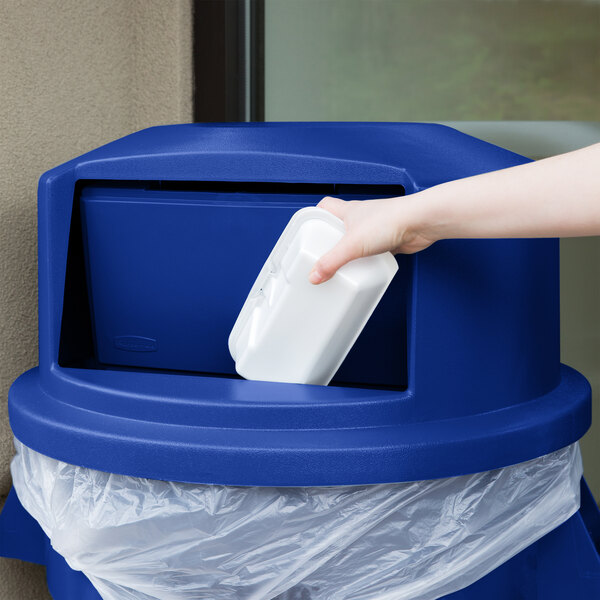 A hand putting a plastic bag into a blue Rubbermaid BRUTE dome top lid on a blue trash can.