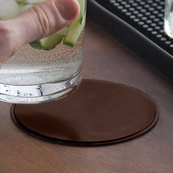 A hand holding a glass of water with cucumber on a brown round vinyl coaster.