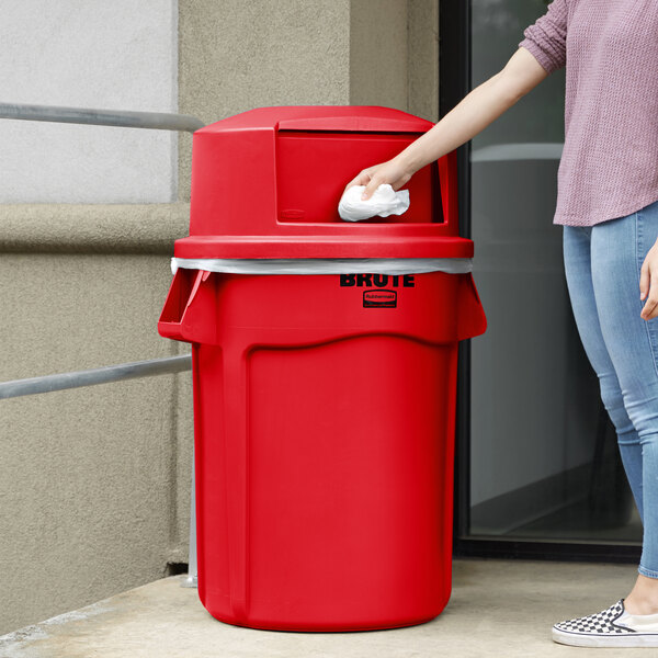 A woman throwing a white tissue into a red Rubbermaid trash can.