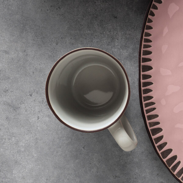 A white stoneware mug with a brown rim next to a pink plate on a gray table.