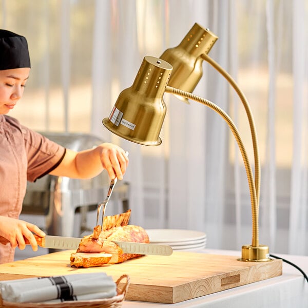 A woman using a knife to cut meat on a wooden cutting board under a gold heat lamp.