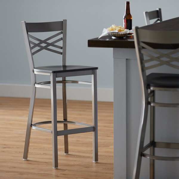 A Lancaster Table & Seating cross back bar stool with a black wood seat next to a table.