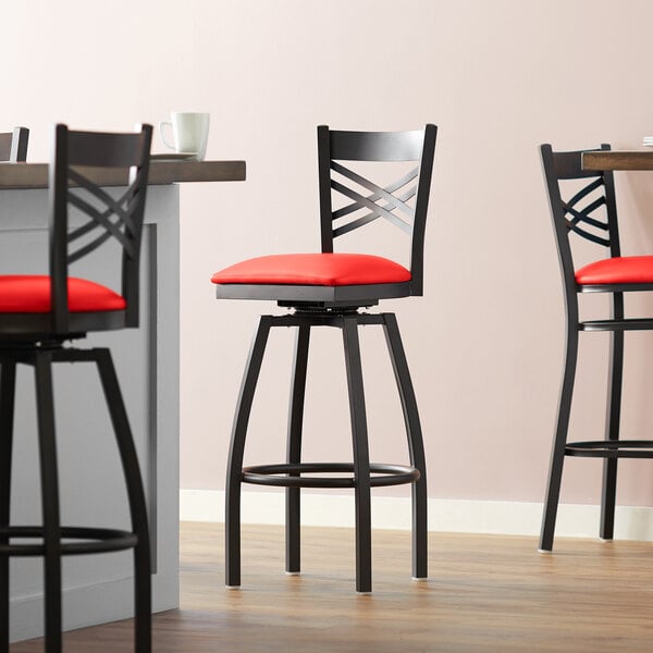 A group of three Lancaster Table & Seating black cross back swivel bar stools with red padded seats.