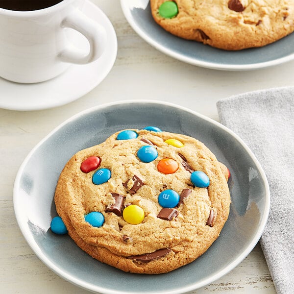 A plate with two David's Cookies M&M's Chocolate Chunk cookies with candy on top on a table with a white mug of coffee.