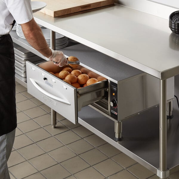 A man using a ServIt single freestanding drawer warmer to hold a tray of doughnuts on a counter.