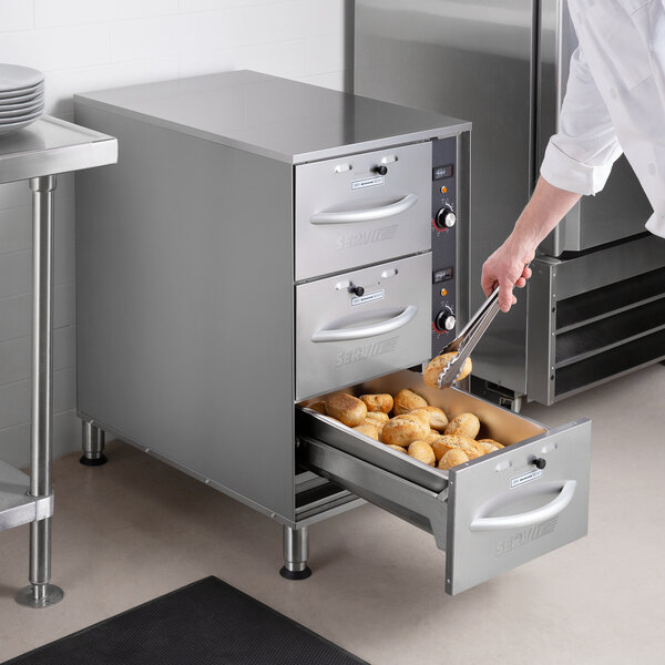 A chef using tongs to put food in a ServIt narrow freestanding drawer warmer.