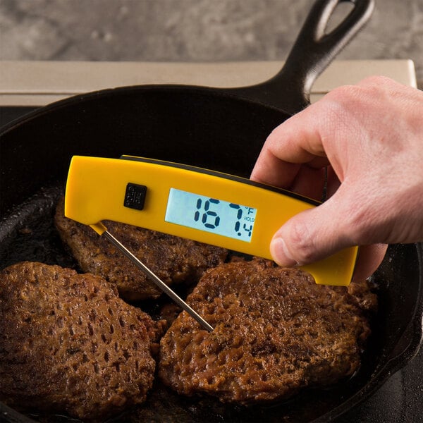 A hand holding a Taylor yellow digital thermometer over meat.