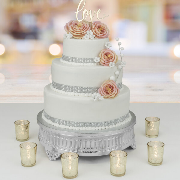 A white wedding cake with pink flowers on a Tabletop Classics by Walco nickel-plated cake stand on a table in a bakery display.