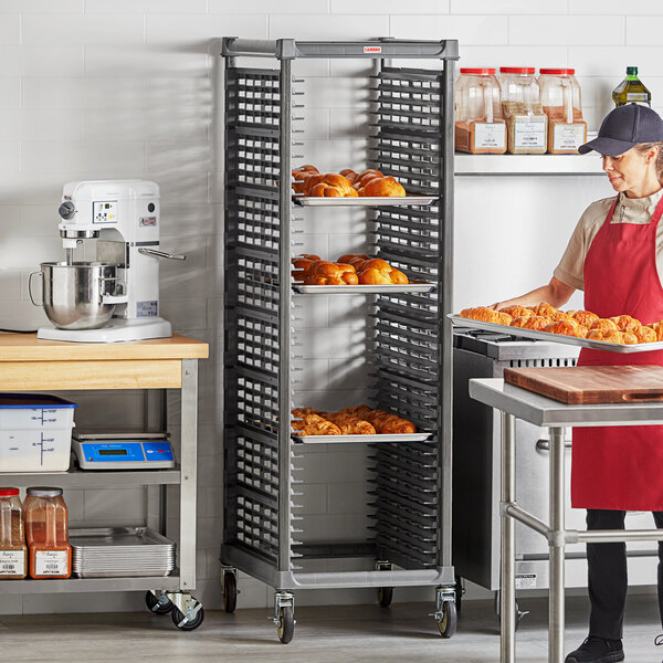 A woman in a bakery standing in front of a Cambro sheet pan rack holding a tray of doughnuts.