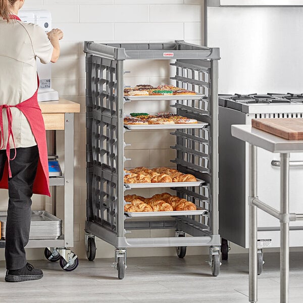 A woman in a red apron standing in a school kitchen with a Cambro sheet pan rack full of pastries.