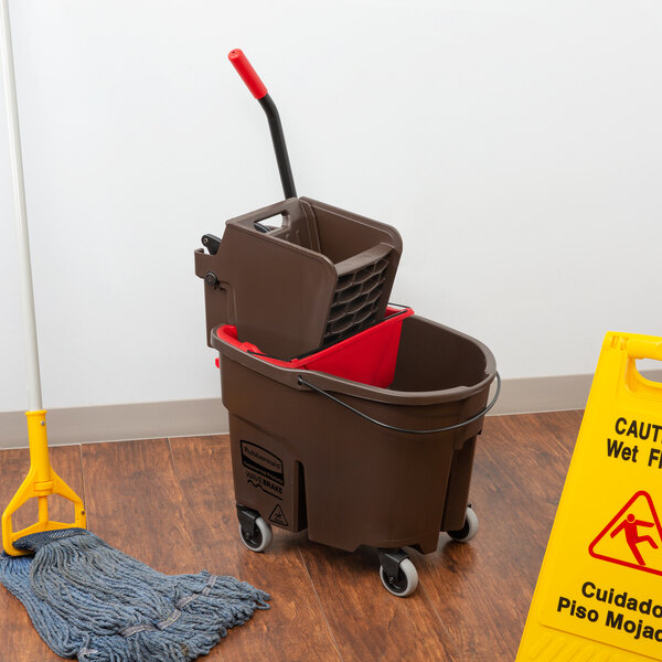 A Rubbermaid WaveBrake mop bucket and wringer on the floor next to a yellow caution sign.