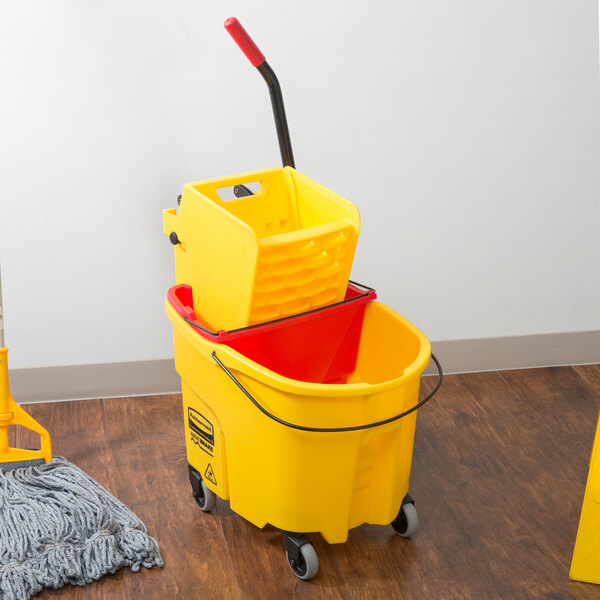 A Rubbermaid yellow mop bucket with a red dirty water bucket next to a mop on a wood floor.