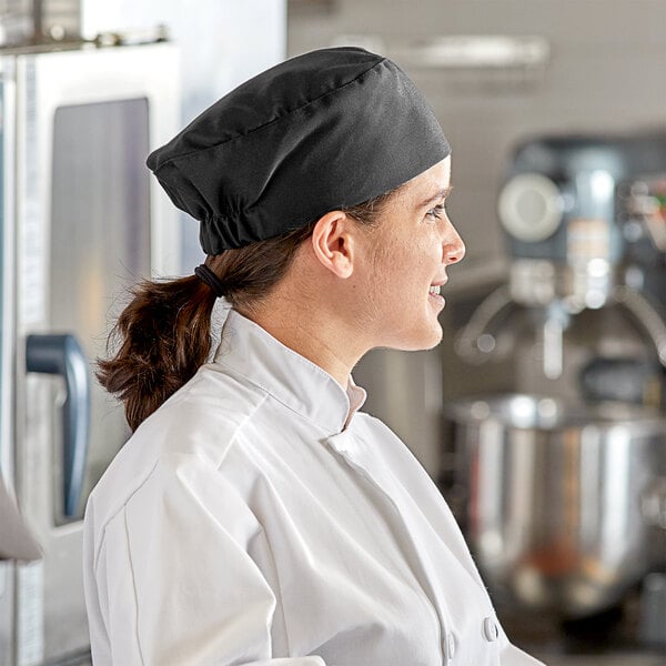 A woman in a black Choice chef's hat and uniform holding a tray of food on a counter in a professional kitchen.