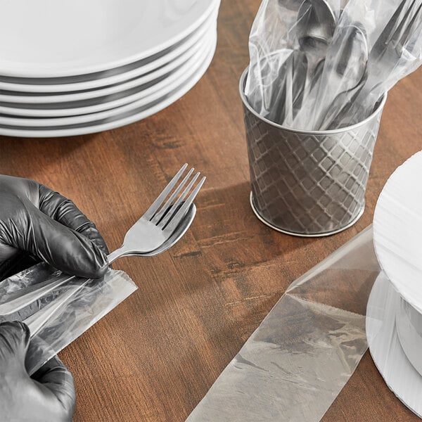 A person in black gloves holding a 3" x 11" plastic bag with a fork and knife inside.