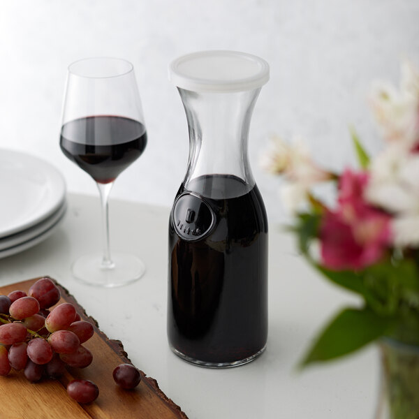 A Libbey wine decanter filled with red wine on a table next to a bottle of wine and grapes.
