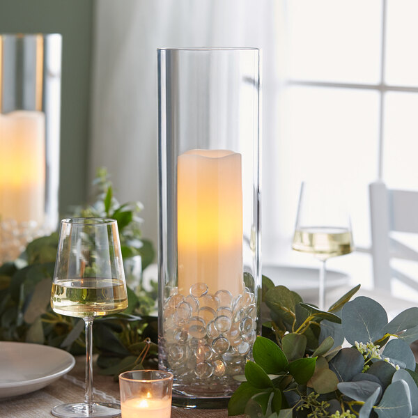 A candle in a clear glass vase with marbles and a glass of white wine on a table.