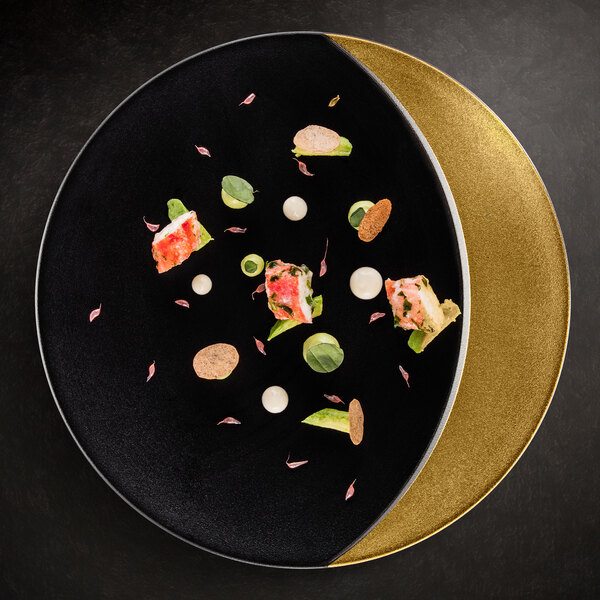 A black RAK Porcelain flat plate with gold accents holding food on a table.