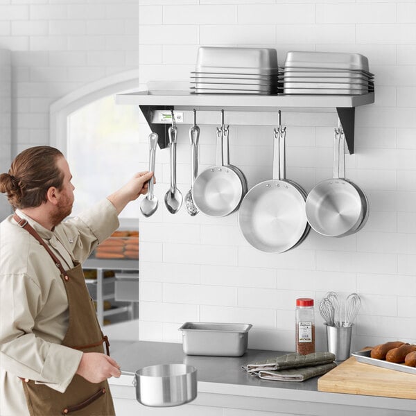 A man in an apron standing in front of a Regency stainless steel wall mounted pot rack with pots and pans hanging from it.