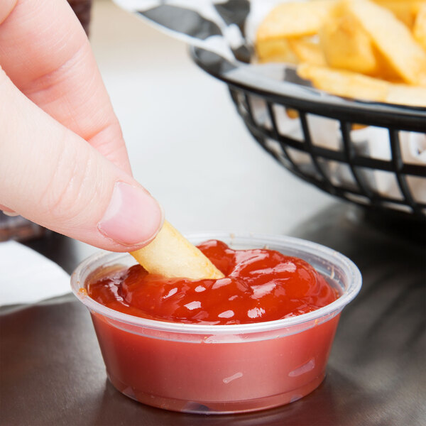 A person dipping a french fry into a Dart plastic souffle cup of ketchup.