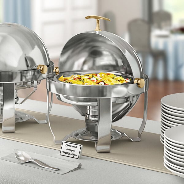 An Acopa Supreme gold-trimmed round roll top chafer on a buffet table with white plates and silver spoons.