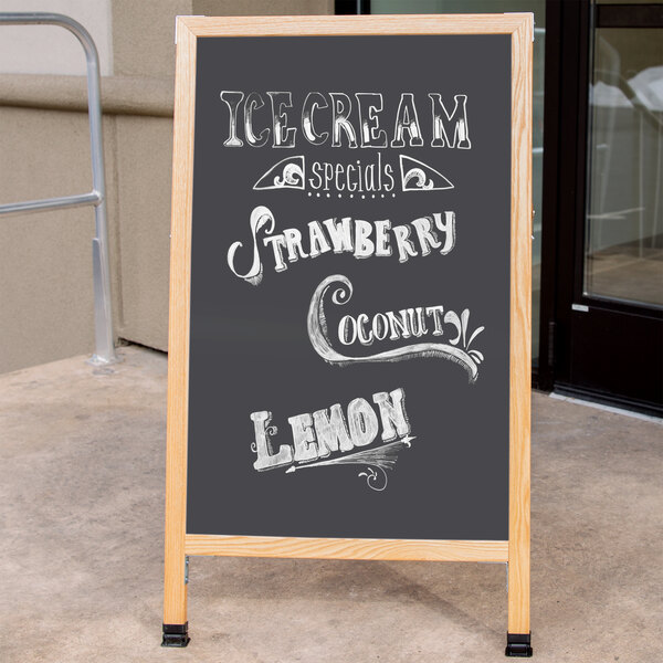 An Aarco oak A-frame chalkboard sign on a sidewalk with white writing advertising ice cream, strawberry, lemon and cream.