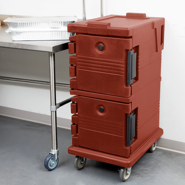 A brick red Cambro Ultra Camcart food pan carrier on wheels next to a table.