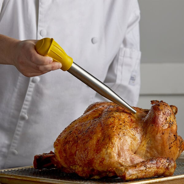 A person using a Fox Run stainless steel turkey baster with a yellow handle to baste a turkey on a metal rack.