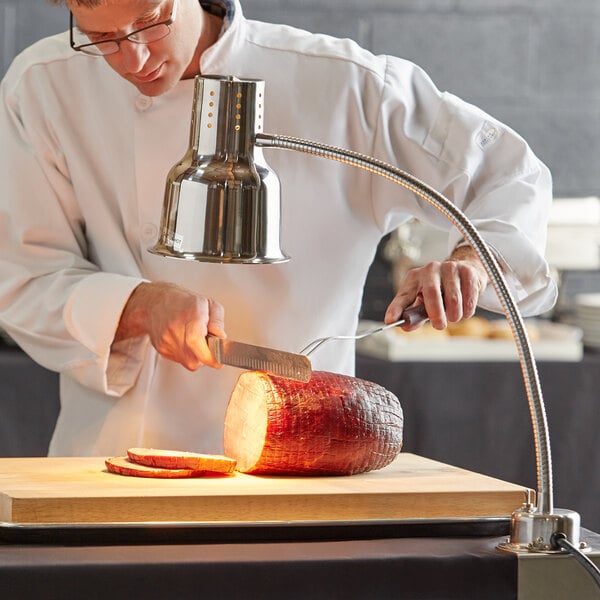 A man in a chef's uniform using an Avantco stainless steel heat lamp to cut a piece of meat.