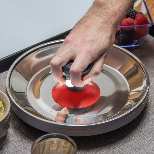 A hand using a black and red Dinex Palm Grip lifter to remove a wax base from a metal container on a silver plate.