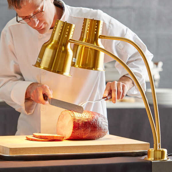 A man in a white coat uses a knife to cut a piece of meat under a gold Avantco heat lamp.