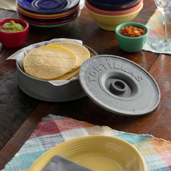 A stack of colorful plates and a gray tortilla warmer with tortillas inside.
