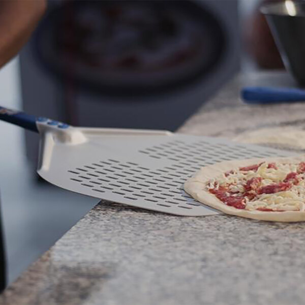 A pizza being put on a square perforated pizza peel with a long handle.