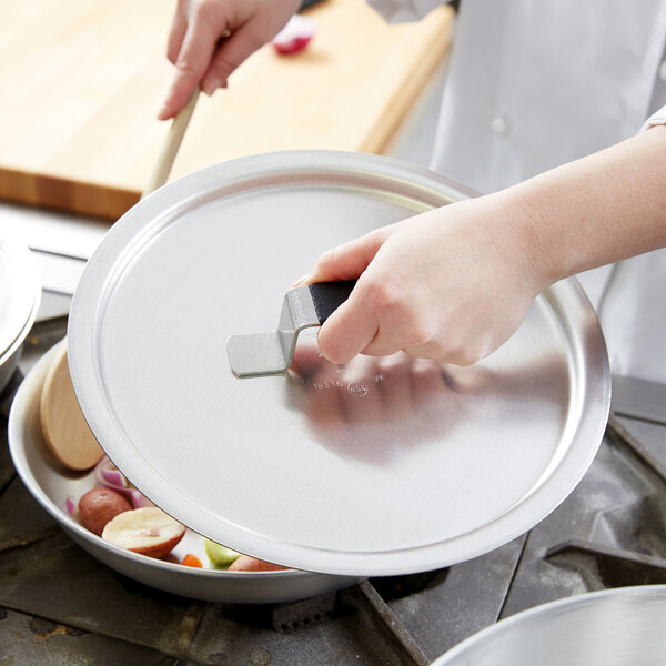 A person holding a Vollrath metal pot cover over a pan with a spatula.