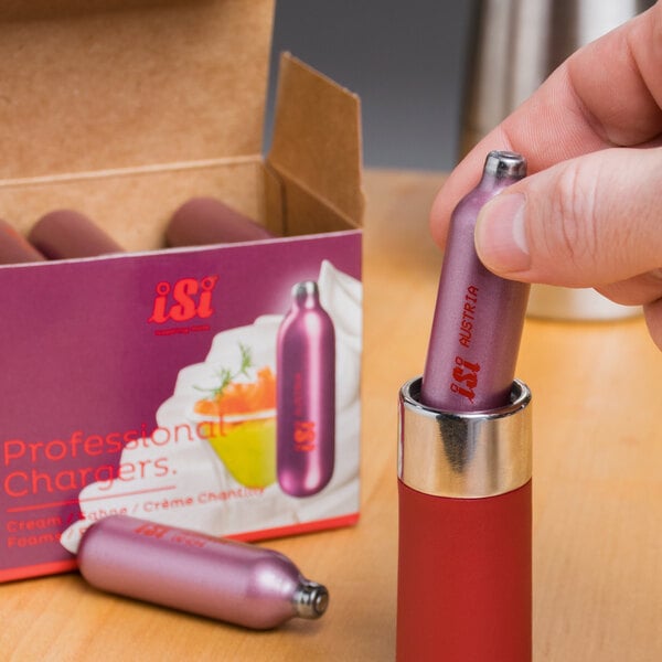 A hand holding a small purple iSi N2O cream charger in front of a box.