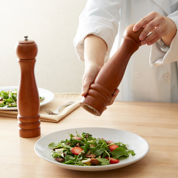 A person in a white coat using an Acopa wooden pepper mill to add pepper to a salad.