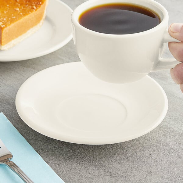 An Acopa ivory saucer under a cup of coffee held by a hand.