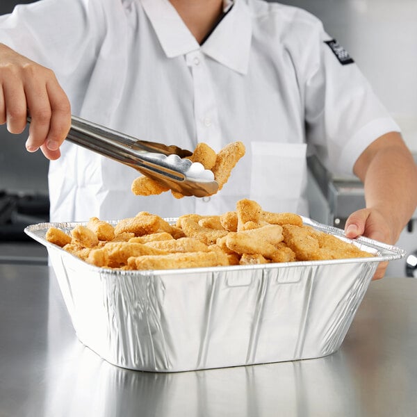 A woman using metal tongs to put chicken nuggets in a Western Plastics foil tray.