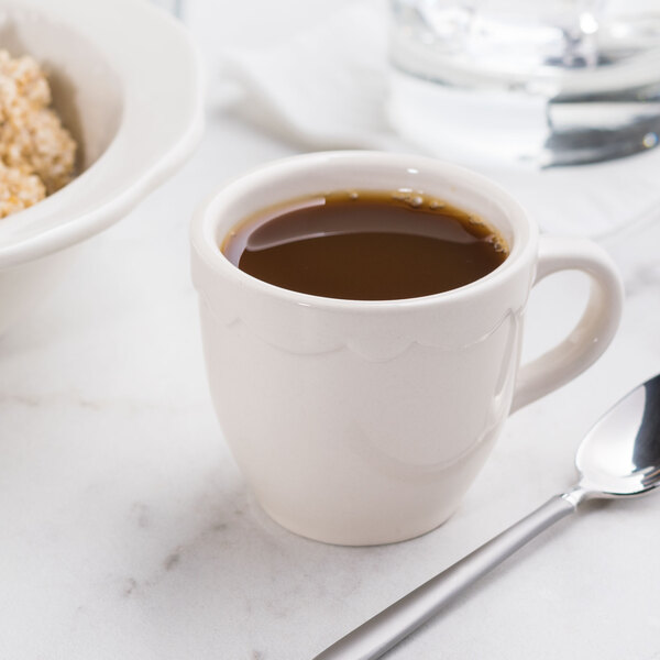 A CAC ivory scalloped edge espresso cup filled with brown liquid with a spoon.