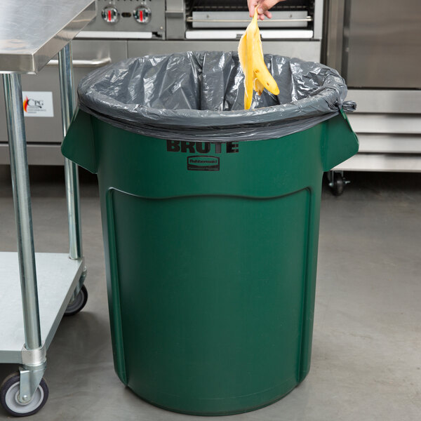A hand in a yellow glove holding a banana in a green Rubbermaid Brute garbage can.
