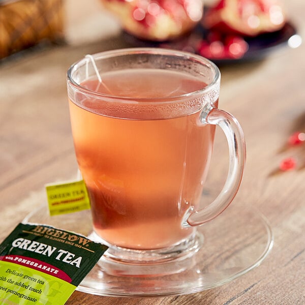 A glass cup of Bigelow green tea with a pomegranate tea bag on a table.