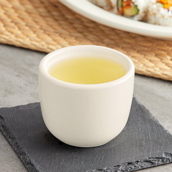 An Acopa ivory sake tea cup filled with yellow liquid on a black plate.